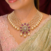 A close-up of a model wearing an ornate Indian-style gold necklace with white faux pearls and a central floral motif set with pink gemstones and a clear central stone, paired with a light green and pink saree.