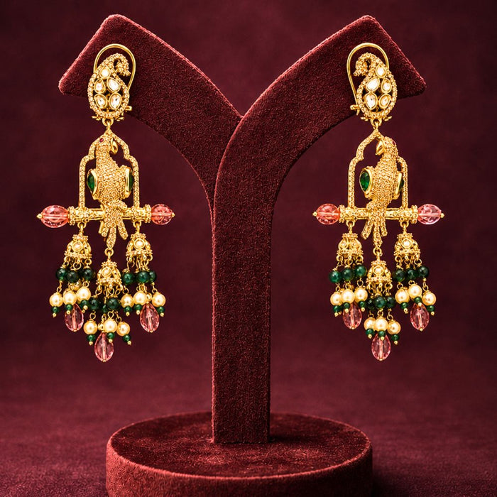 A pair of ornate gold-toned Indian-style dangle earrings with green and pink stones and pearl-like beads, displayed on a dark red velvet jewelry stand against a matching dark red background.