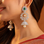 A close-up lifestyle shot of a woman wearing a pair of intricate gold-plated dangle earrings. The earrings feature a green oval-cut stone at the top, surrounded by clear pavé stones, with two cascading circular motifs and a final bell-shaped jhumka adorned with a single white pearl.