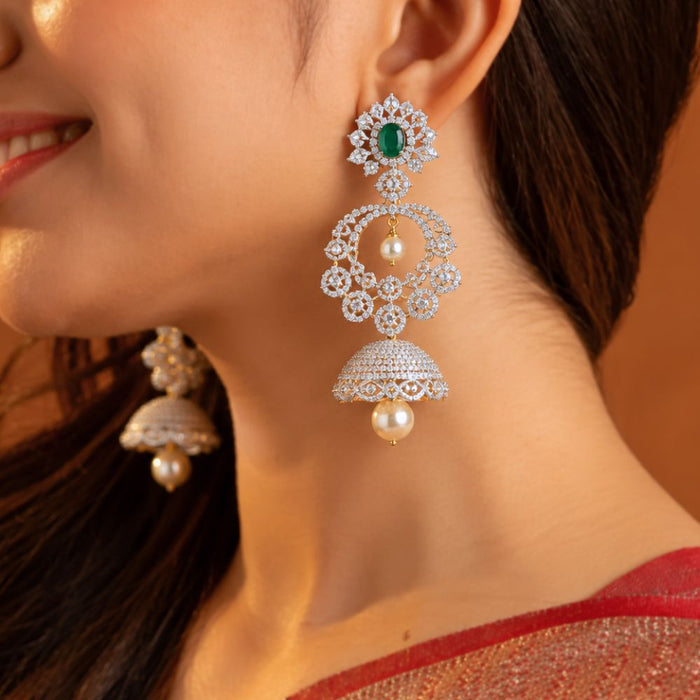 A close-up lifestyle shot of a woman wearing a pair of intricate gold-plated dangle earrings. The earrings feature a green oval-cut stone at the top, surrounded by clear pavé stones, with two cascading circular motifs and a final bell-shaped jhumka adorned with a single white pearl.