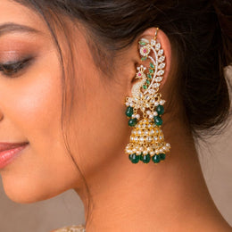 A close-up of a woman's ear wearing a large, ornate gold-toned Jhumka earring decorated with white stones, green gemstones, and small pink accents, against a soft beige background.