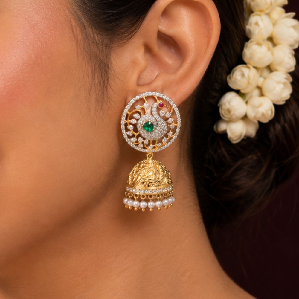 A close-up of a woman's ear wearing a traditional Indian gold and silver jhumka earring with a green stone and white stones, next to white flowers in her hair.