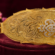 A close-up of an ornate gold belt buckle with intricate floral engravings and a diamond-encrusted floral motif, displayed on a deep burgundy velvet surface.South Indian Bridal Waist Belt with Green Stones & CZ Work