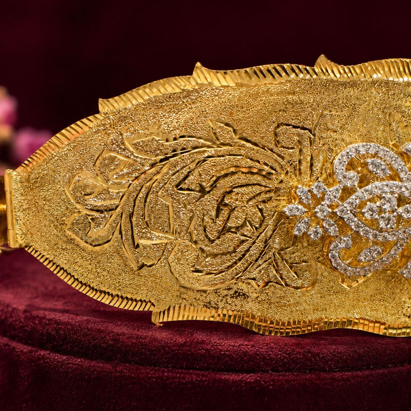A close-up of an ornate gold belt buckle with intricate floral engravings and a diamond-encrusted floral motif, displayed on a deep burgundy velvet surface.South Indian Bridal Waist Belt with Green Stones & CZ Work