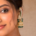 A close-up of a woman wearing a large, ornate gold-plated jhumka earring decorated with green stones, white faux pearls, and small red stones, against a neutral beige background.