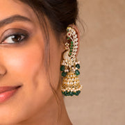 A close-up of a woman wearing a large, ornate gold-plated jhumka earring decorated with green stones, white faux pearls, and small red stones, against a neutral beige background.