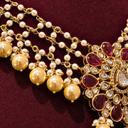 Gold-toned traditional Indian jewelry featuring a necklace with dangling pearl-like beads and a heavily embellished peacock motif set with deep red teardrop stones and clear crystals, displayed on a dark burgundy fabric background.