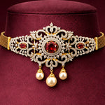 An ornate gold and silver-toned choker necklace displayed on a dark burgundy velvet jewelry bust. The choker features a central round red gemstone surrounded by silver pavé, flanked by two smaller red gemstones, with three dangling cream pearl beads below.