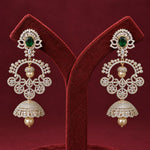 A pair of ornate gold-toned dangle earrings with green stones and pearl-like beads, displayed on a red velvet jewelry stand against a dark red background.
