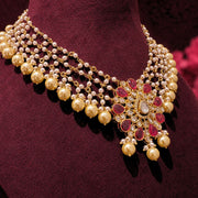 An ornate gold-plated Indian style statement necklace with faux pearls, red stones, and clear crystals, displayed on a dark burgundy velvet jewelry bust.