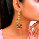 A close-up lifestyle shot of a woman wearing a large, ornate gold-toned Indian chandelier earring decorated with green and pink stones.
