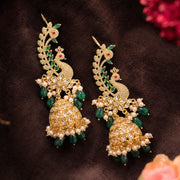 A pair of ornate gold-toned traditional Indian jhumka earrings decorated with green stone peacock motifs, white pearl beads, and small red gemstone accents, displayed on a dark brown textured fabric background.