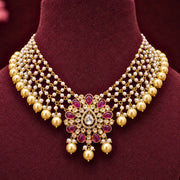 An ornate Indian style gold-plated necklace with multiple strands of white faux pearls and a central floral pendant featuring red gemstones and a clear central stone, displayed on a dark burgundy velvet jewelry bust.