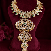 An ornate gold-toned traditional necklace with green and white stones, displayed on a dark burgundy velvet jewelry bust.