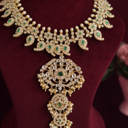 An ornate gold-toned traditional necklace with intricate diamond-like stones and green stone accents, displayed on a dark red velvet jewelry bust.