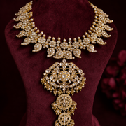 An ornate gold-toned statement necklace with intricate crystal and pearl-like embellishments, displayed on a dark burgundy velvet jewelry bust against a dark red background.