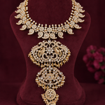 An ornate gold-toned jewelry necklace with intricate rhinestone and crystal embellishments, displayed on a dark burgundy velvet bust. The necklace features a wide leaf-patterned collar and a large, symmetrical pendant with peacock motifs and dangling crystal drops.