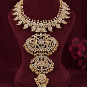 An ornate gold-toned jewelry necklace with intricate rhinestone and crystal embellishments, displayed on a dark burgundy velvet bust. The necklace features a wide leaf-patterned collar and a large, symmetrical pendant with peacock motifs and dangling crystal drops.