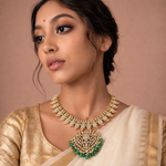 A woman wearing an ornate gold and green traditional necklace with a large pendant, paired with a cream-colored sari. The necklace features intricate gold detailing, clear stones, and green bead accents, set against a solid warm brown background.