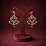 A pair of ornate gold-toned dangle earrings with green teardrop gemstones, displayed on a dark brown velvet jewelry stand against a matching dark brown background.