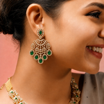 A close-up lifestyle shot of a woman wearing a gold-toned chandelier earring with green emerald-cut stones and a matching necklace against a soft pink background.