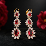 Long champagne-gold statement earrings with graduated pear-shaped ruby-red stones and marquise-cut white floral halos. Ideal for weddings, receptions and festive occasions.