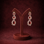 A pair of long drop earrings with red gemstones and clear crystal-like floral settings, displayed on a brown velvet jewelry stand against a dark burgundy background.
