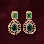 A pair of gold drop earrings featuring emerald-cut green stones at the top and teardrop-cut green stones in the center, surrounded by small clear stones, set against a dark burgundy background.
