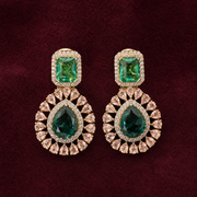 A pair of gold drop earrings featuring emerald-cut green stones at the top and teardrop-cut green stones in the center, surrounded by small clear stones, set against a dark burgundy background.