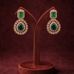 A pair of gold and emerald green gemstone drop earrings displayed on a dark brown velvet jewelry stand against a dark burgundy background.