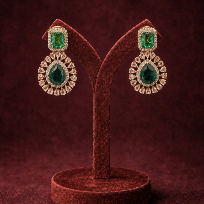 A pair of gold and emerald green gemstone drop earrings displayed on a dark brown velvet jewelry stand against a dark burgundy background.