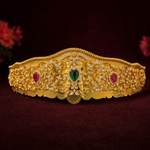 An ornate gold crown with intricate filigree detailing, set with a central green teardrop gemstone and two red oval gemstones, displayed on a dark burgundy background.