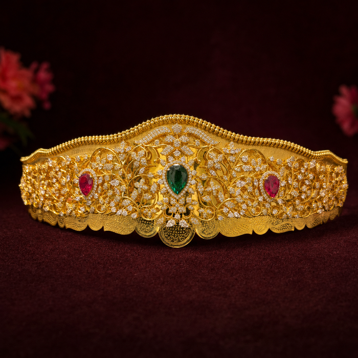 An ornate gold crown with intricate filigree detailing, set with a central green teardrop gemstone and two red oval gemstones, displayed on a dark burgundy background.