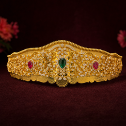 An ornate gold crown with intricate filigree detailing, set with a central green teardrop gemstone and two red oval gemstones, displayed on a dark burgundy background.