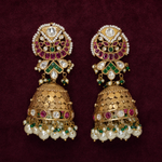 A pair of ornate Indian jhumka earrings in gold-plated metal, embellished with white pearls, green and pink stones, and clear crystals, displayed on a dark burgundy velvet background.