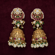A pair of ornate Indian jhumka earrings in gold-plated metal, embellished with white pearls, green and pink stones, and clear crystals, displayed on a dark burgundy velvet background.