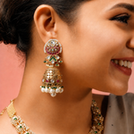 A close-up lifestyle shot of a woman wearing an ornate gold-toned Indian jhumka earring with white pearls and red, green, and white stones, paired with a matching gold necklace against a soft pink background.
