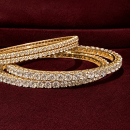 Three gold bangles adorned with rows of sparkling clear stones, arranged on a deep burgundy velvet background.