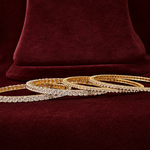 Two gold bangle bracelets, one with clear pavé stones and one with small clear stones and gold accents, displayed on a deep burgundy velvet surface.