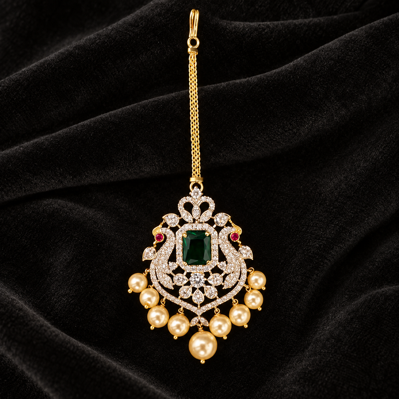 An ornate gold-toned Indian maang tikka pendant with a central green emerald-cut stone, surrounded by clear pavé stones, small red gem accents, and dangling pearl-like beads, displayed on a black velvet background.