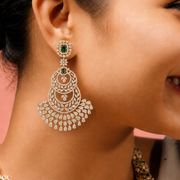 A close-up lifestyle shot of a woman wearing an ornate gold and diamond chandelier earring with green stone accents, along with a matching necklace, set against a soft pink background.