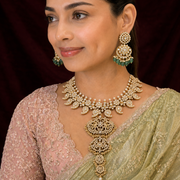 A woman wearing an ornate gold-toned traditional necklace with matching drop earrings, styled with a light green saree and a pink embroidered blouse against a dark maroon backdrop.