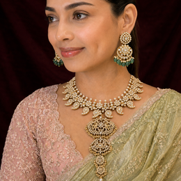 A woman wearing an ornate gold-toned traditional necklace with matching drop earrings, styled with a light green saree and a pink embroidered blouse against a dark maroon backdrop.