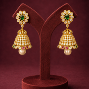 A pair of ornate gold-toned jhumka earrings displayed on a dark red velvet jewelry stand against a deep burgundy background. Each earring features a floral stud with a green stone center and clear stones, a bell-shaped drop covered in clear stones and gold detailing, and a dangling pearl.