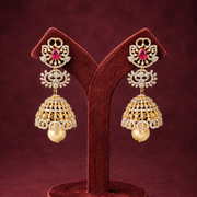 A pair of ornate gold-toned Indian jhumka earrings with red stones and white stones, displayed on a dark red velvet jewelry stand against a dark red background.