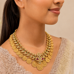 A close-up of a woman wearing an ornate gold coin necklace with green and red stone accents, paired with a light beige embroidered top against a soft beige background.