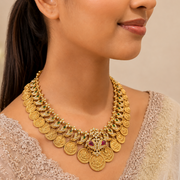 A close-up of a woman wearing an ornate gold coin necklace with green and red stone accents, paired with a light beige embroidered top against a soft beige background.