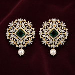 A pair of ornate gold and diamond drop earrings with a central green emerald-cut stone and a dangling white pearl, displayed on a dark burgundy velvet background.