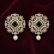 A pair of ornate gold and diamond drop earrings with a central green emerald-cut stone and a dangling white pearl, displayed on a dark burgundy velvet background.