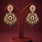 A pair of ornate gold-toned Indian-style jhumka earrings with green and red stones, displayed on a dark red velvet jewelry stand against a matching dark red background.